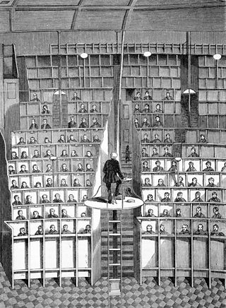 A drawing of a large room where a priest stands on a platform facing ten rows of prisoners sitting in tiers of separated wooden cubicles.